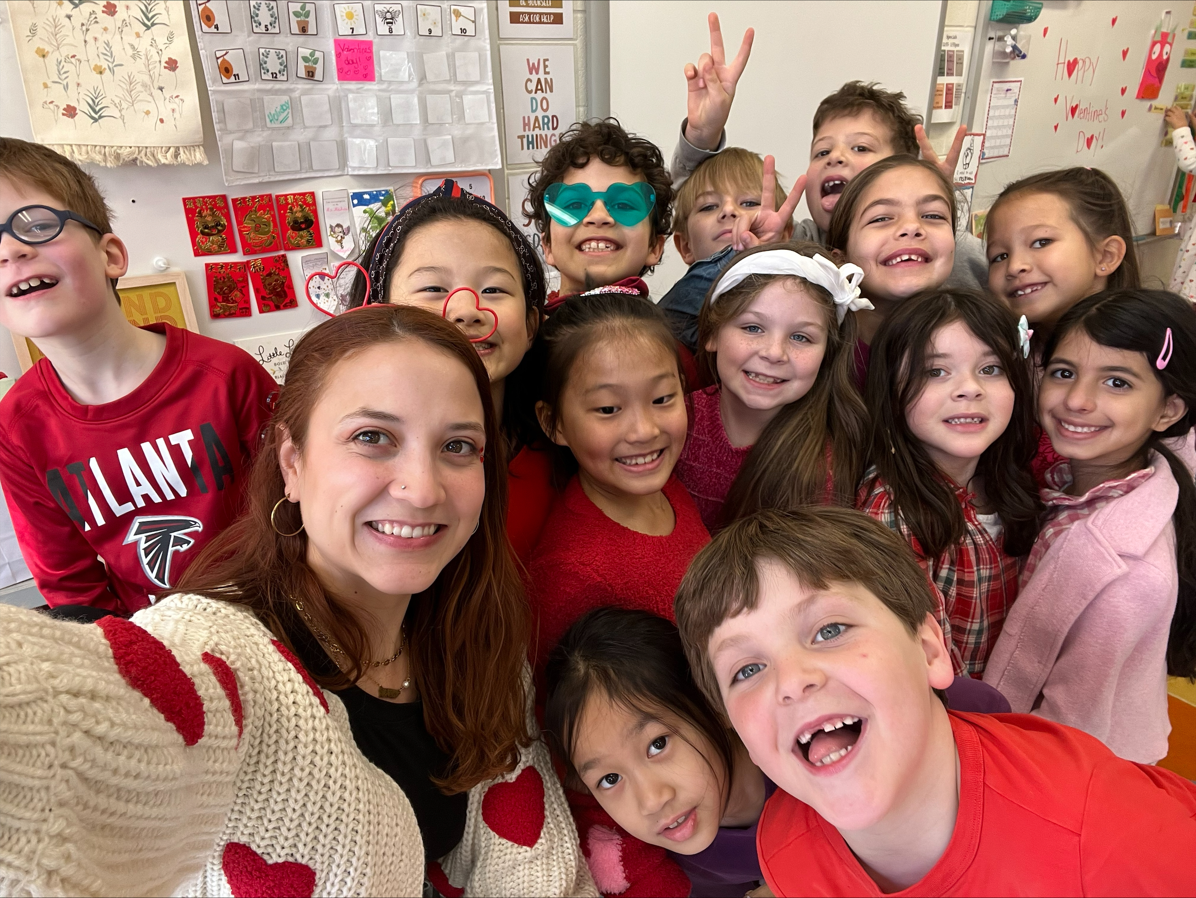 self portrait of Ms. medina with several students on valentines day. They're all smling and wearing red, pink, and hearts. the background shows the front of their second grade classroom.
