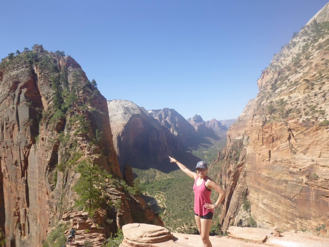 Ms. choi stands on a hiking trail in Zion National Park pointing at the summit objective. she wears black shorts, a sleeveless athletic top, and a grey baseball cap. the terrain is rocky with patches of small green plants. it is a cloudless day above the canyon in the background.