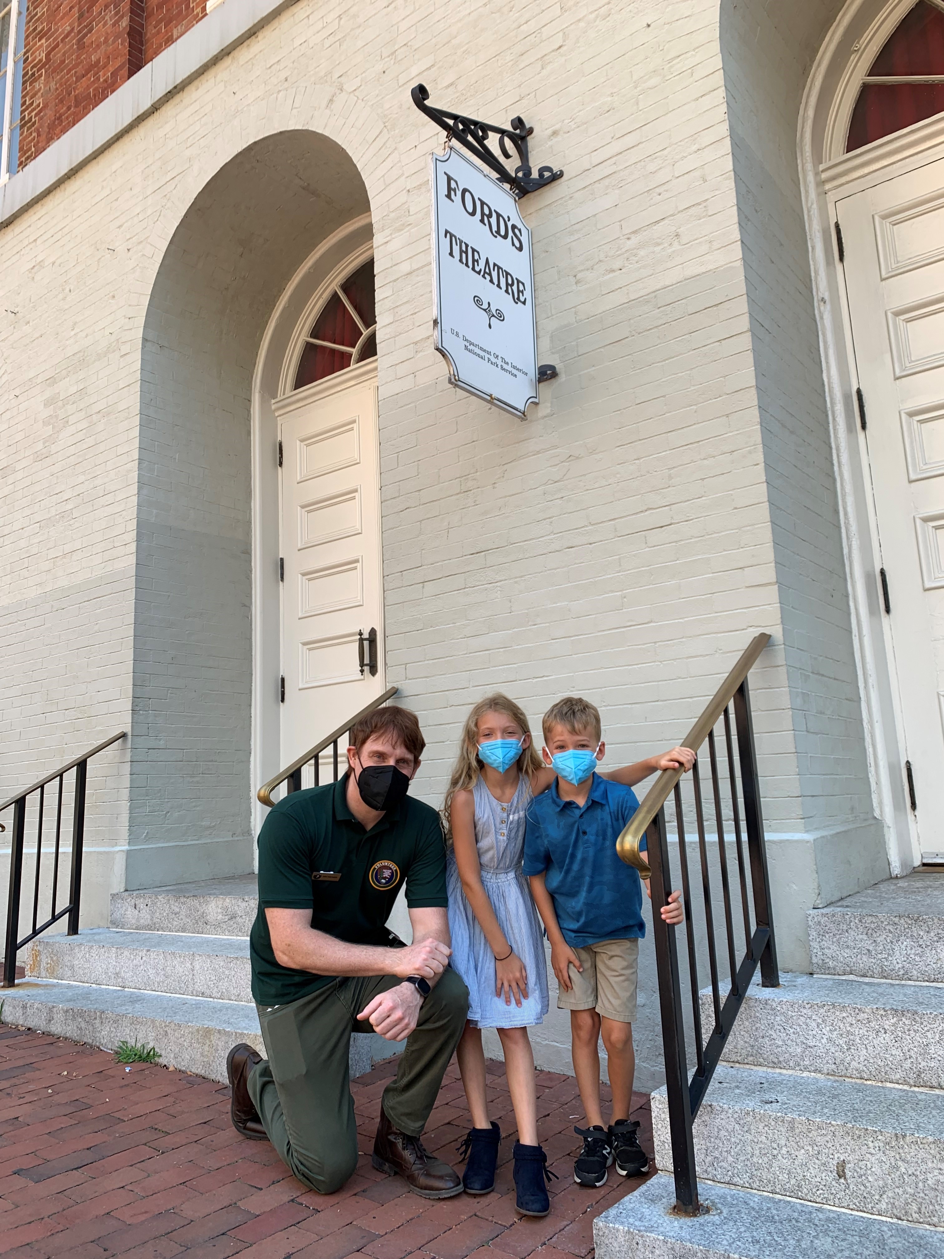 mr. baker poses beneath the ford's theatre sign in his parks service uniform with two students. he wears a forest green polo shirt with an insignia on the left chest, olive green trousers, and brown leather boots.