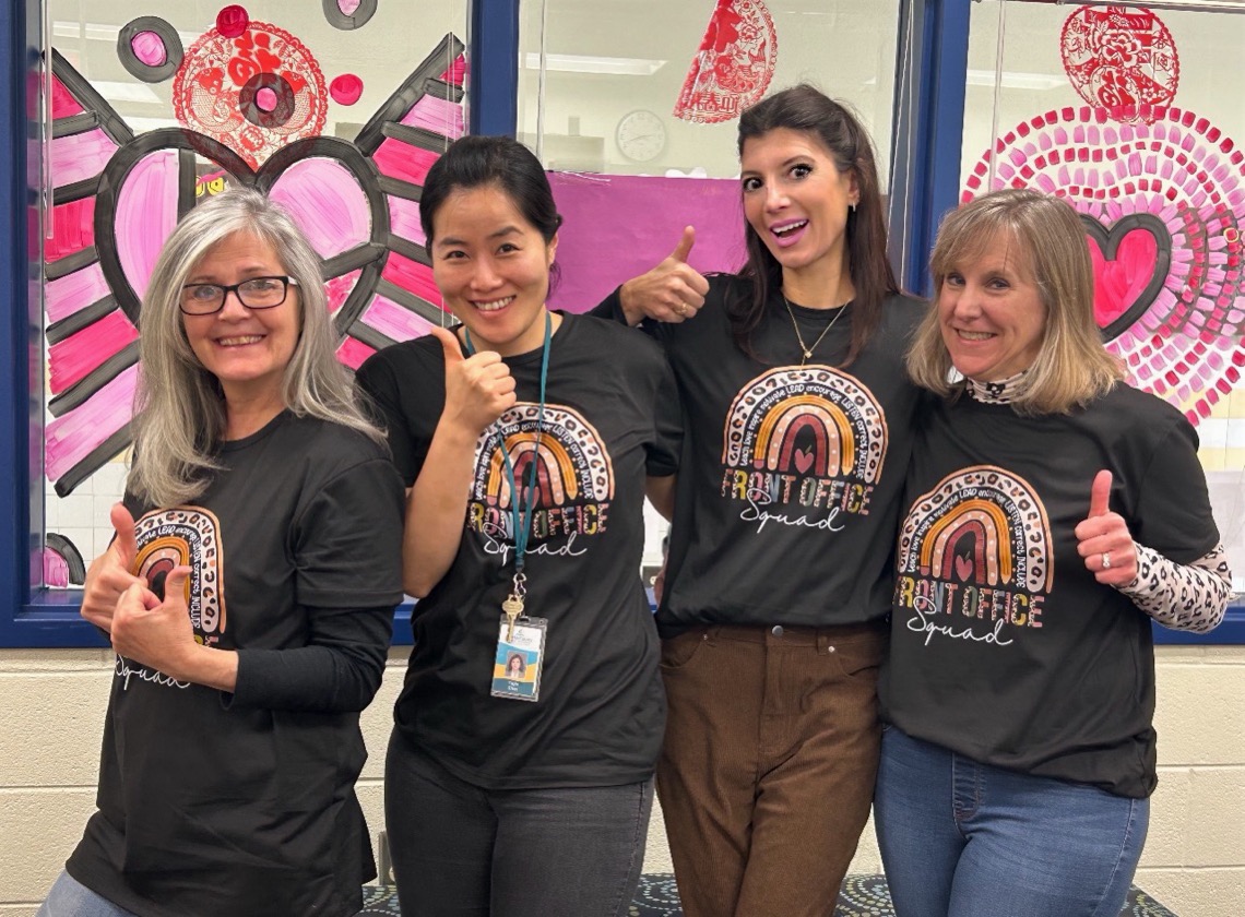 four women from our office staff pose for a picture in front of a pink valentine's design. paulette is second from the right, taller than the rest. wearing a dark grey shirt and brown trousers.