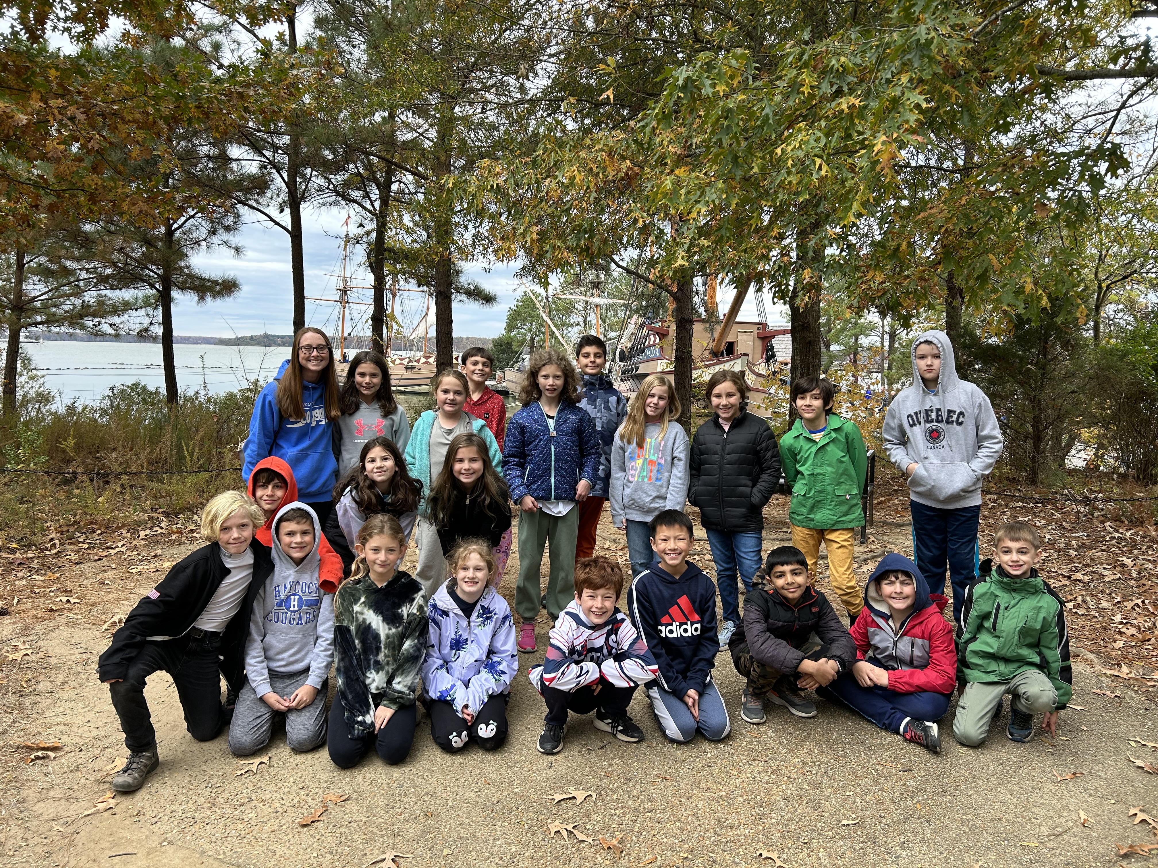 ms. mostoller's class poses for the camera on a field trip. students are dressed in warm clothes for the weather.in the background are trees and a boat on the river near jamestown, virginia.