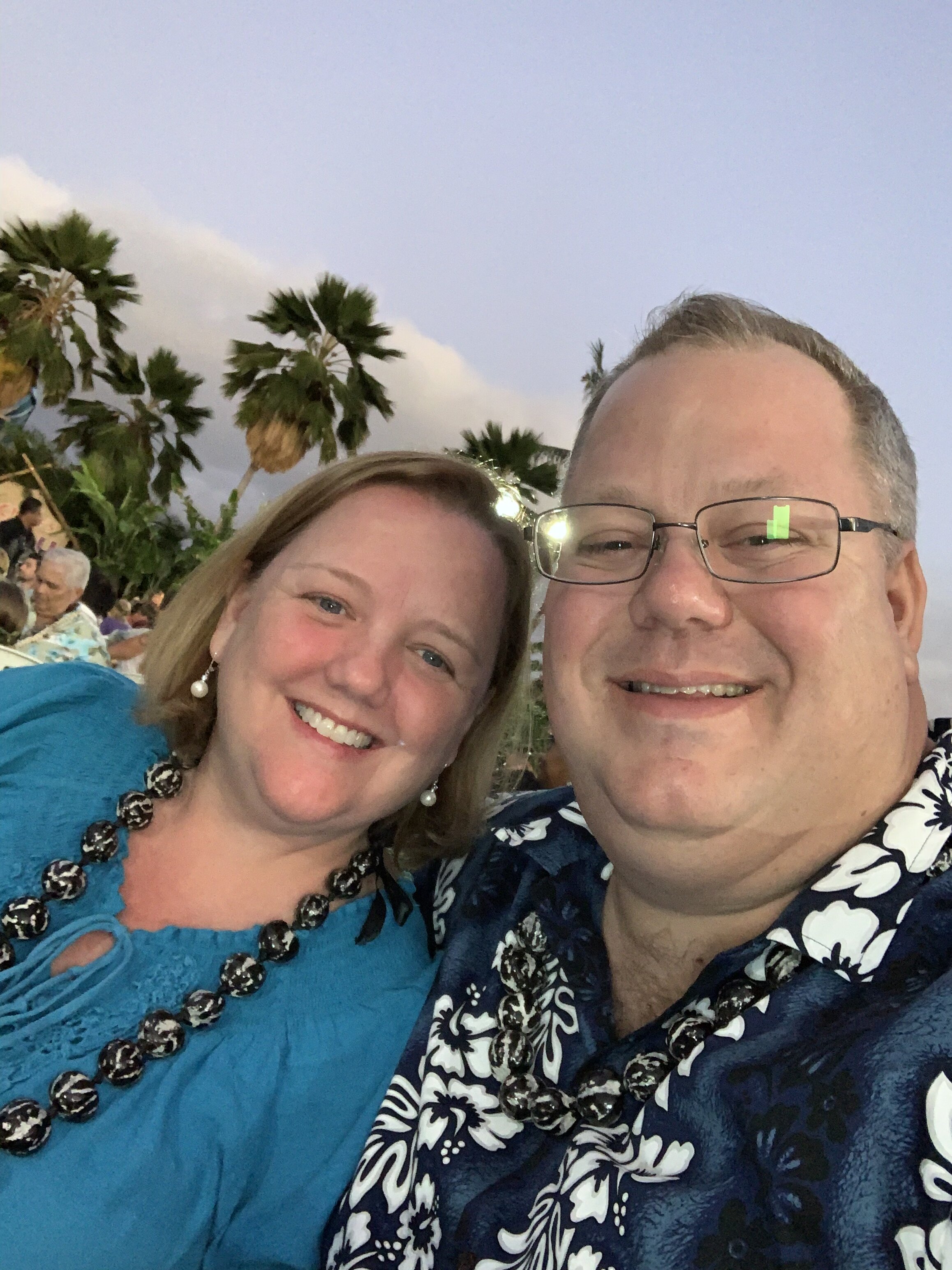 photograph of Mr. Morrow in Hawaii with his wife. She has blond hair and is wearing a teal top. Mr. Morrow is wearing a dark blue floral print shirt and glasses.