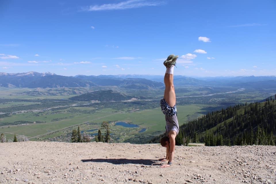 ms. mostoller does a handstand on a hiking trail. in the valley below there are bodies of water, trees, and other mountains.