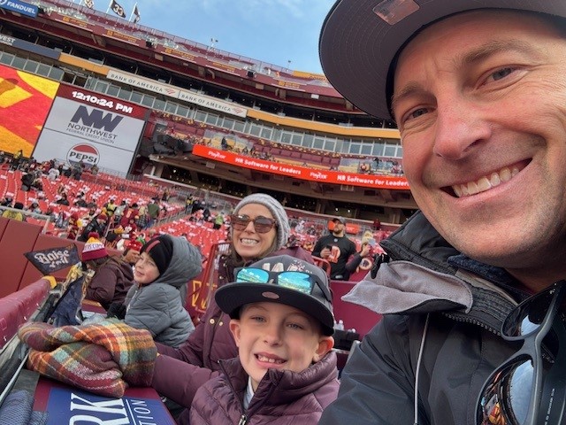 self portrait of mrs. surber with her husband & two sons at a washington commanders football game. they sit near field level with a full stadium behind them.