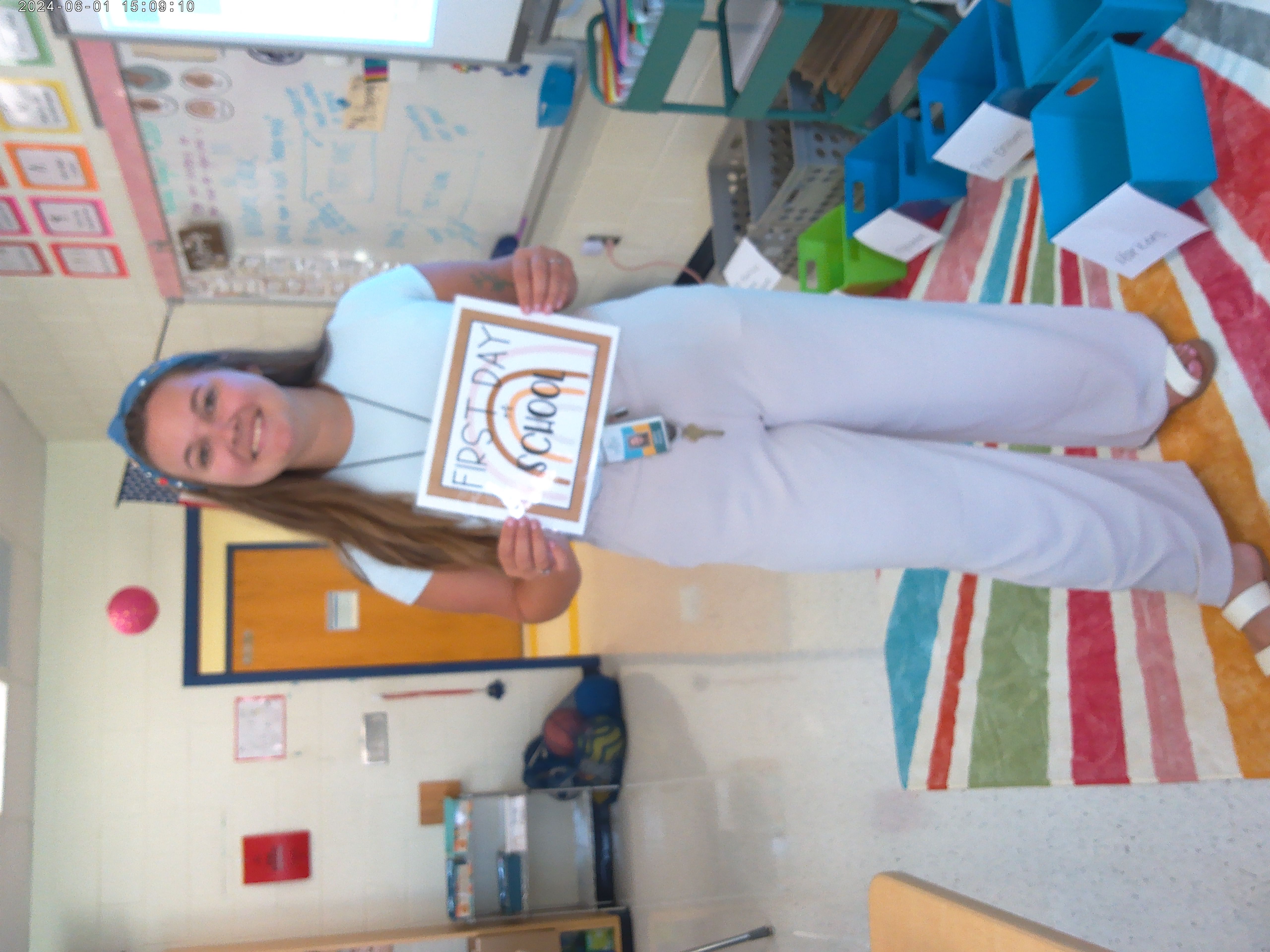 Ms. hopping in her classroom on the first day of school. she stands on a multi-colored striped rug. She wears a blue headband holding back long brown hair. She has a light blue top and flowy linen pants. in her hands is a sign that says "first day of school"