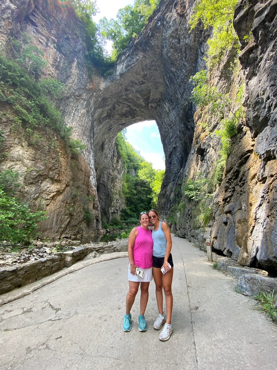 Mrs. Capozzi poses in front of Natural Bridge with one of her daughters. She wears teal sneakers, a white skirt, and a bright pink sleeveless top.