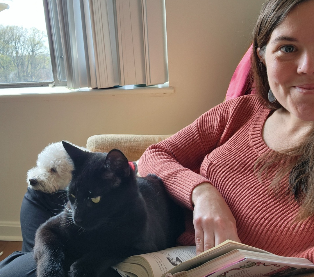 Ms. Padgett poses in front of an open window with a black cat, white dog, and an open book in her lap. she wears a reddish ribbed top.