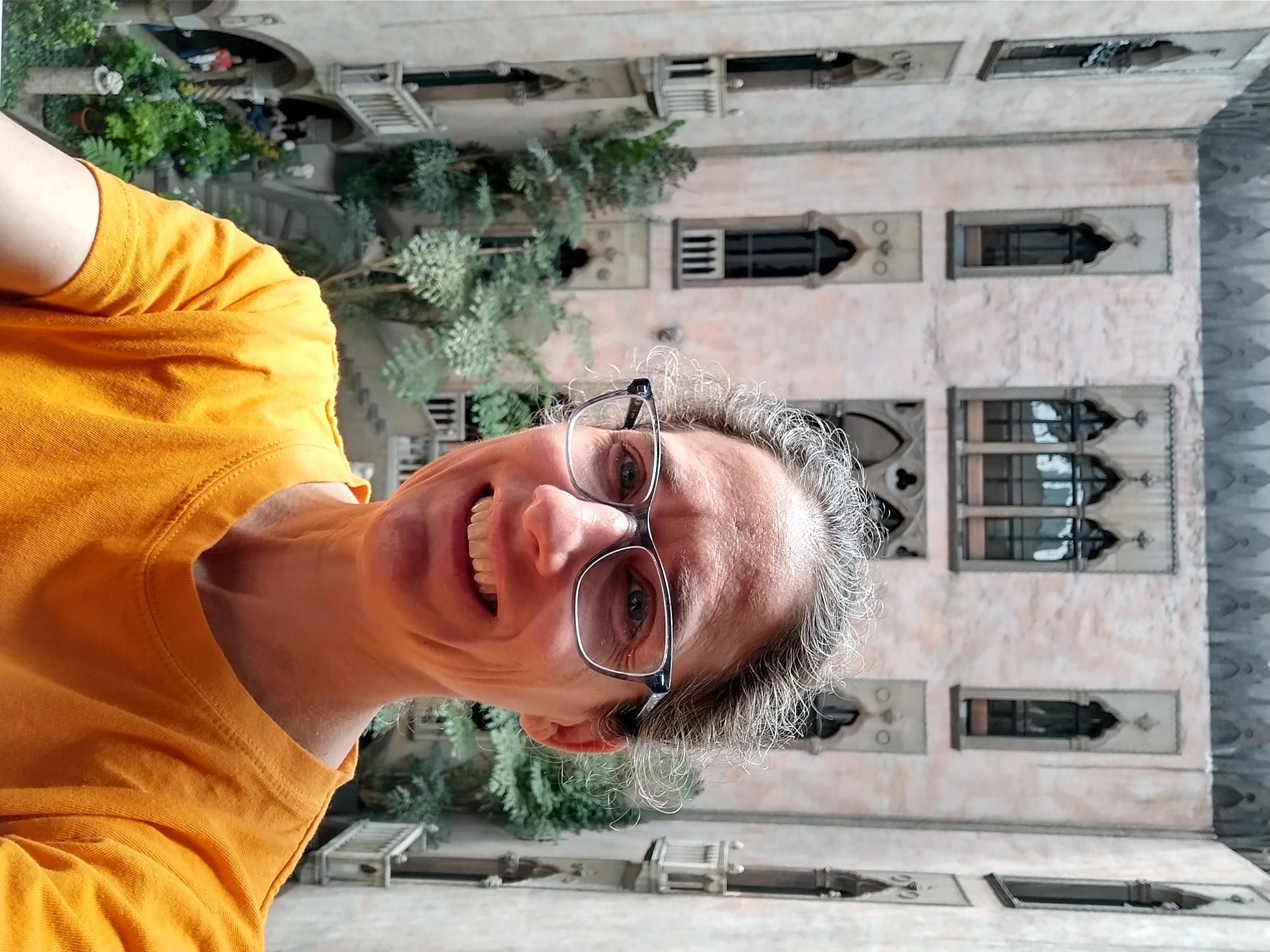 selfie of ms. sklar in glasses and a yellow t-shirt inside of a atrium filled with plants. 