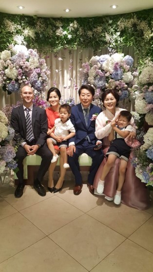 picture of ms. choi with her family. men in picture are wearing dark suits, women are in elegant dresses. boys are wearing matching shorts, white shoes and white shirt. background is framed with colorful flowers in purple, white & green.