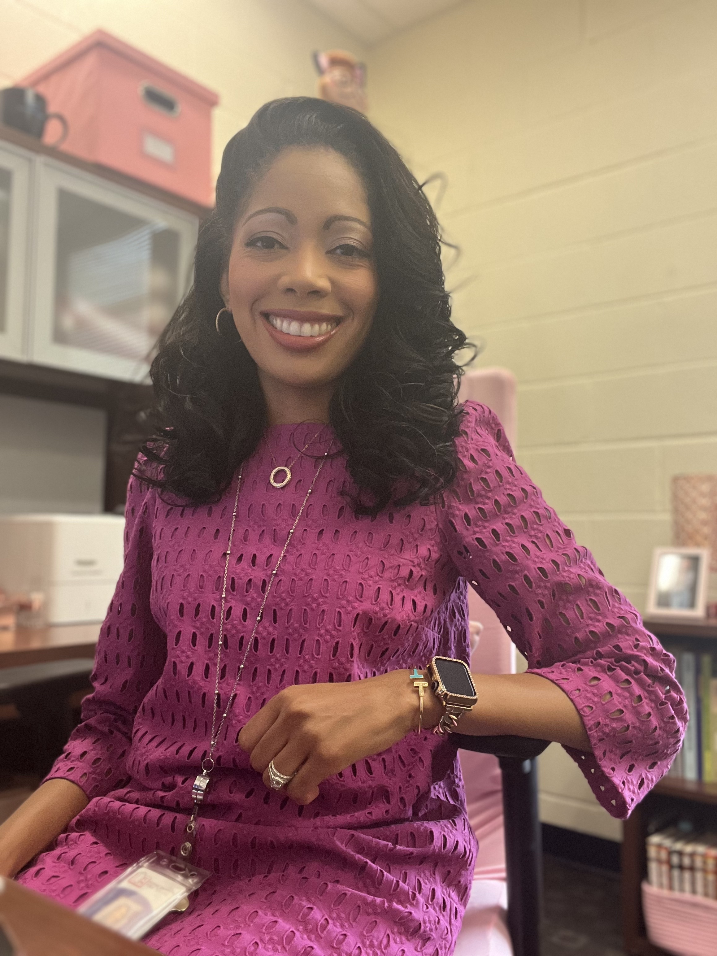 ms. styles poses at her desk in her office. she wears a magenta dress and a friendly smile.