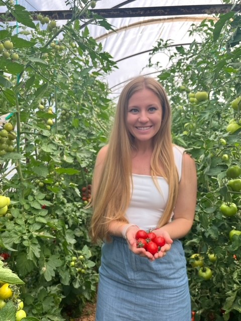 mrs. wisniewski stands in a greenhouse with green tomato plants surrounding her. she holds five ripe tomatoes in her outstretched hands. she's wearing a blue skirt with a white sleeveless blouse covered by her blonde hair.